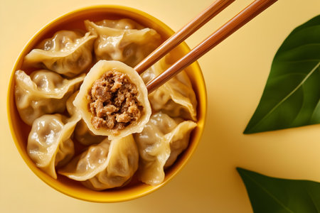 Creative food concept. Pair of chopstick picking up a dumpling, xiao long bao, tangbao, Wonton filled with ground meat from a bowl on yellow background. copy text spaceの写真素材