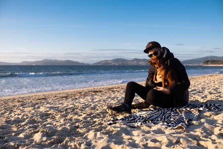 young hispanic man wearing sunglasses and black clothes is hugging his small cute brown dog sitting on the sand at the beach at golder hourの写真素材