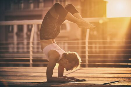 Young woman practicing yoga, standing in Vrischikasana Scorpion pose in the terrace at sunsetの写真素材