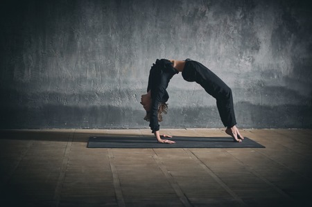 Beautiful woman practices backbend yoga asana Urdhva Dhanurasana - Upward facing bow pose in the dark hallの写真素材
