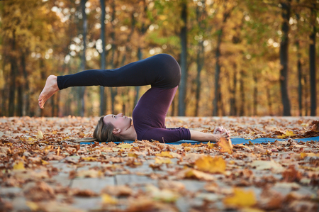 Beautiful young woman practices yoga asana Halasana Plough pose on the wooden deck in the autumn park.の写真素材
