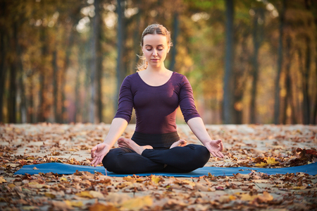 Beautiful young woman meditates in yoga asana Padmasana - Lotus pose on the wooden deck in the autumn park.の写真素材