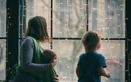 Happy young family, beautiful mother with two children, adorable preschool boy and baby in sling look together through the window to the street.の写真素材