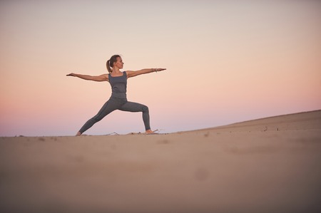 Beautiful young woman practices yoga asana Virabhadrasana 2 - warrior pose 2 in the desert at sunset.の写真素材