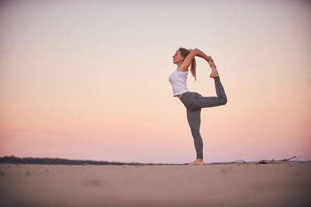 Beautiful young woman practices yoga asana Natarajasana - Lord Of The Dance pose in the desert at sunset.の写真素材