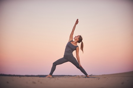 Beautiful young woman practices yoga asana Virabhadrasana 2 - warrior pose 2 in the desert at...