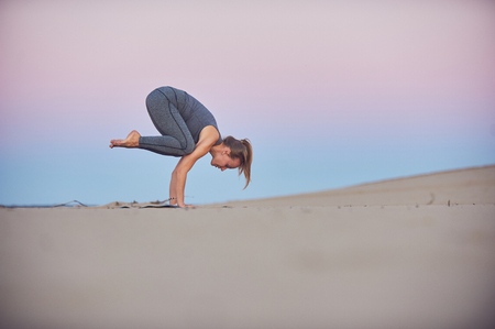 Beautiful young woman practices yoga asana Bakasana - crane pose in the desert at sunset.の写真素材