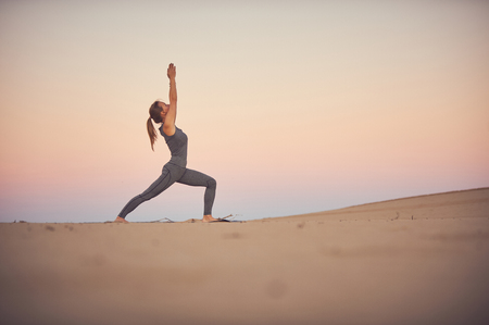 Beautiful young woman practices yoga asana Virabhadrasana 1 - warrior pose 1 in the desert at sunset.の写真素材