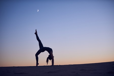 Beautiful young woman practices backbend yoga asana Eka Pada Urdhva Dhanurasana - One Leg Upward facing bow pose in the desert at night.の写真素材