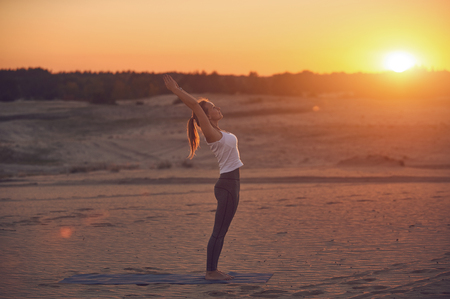 Beautiful young woman practices yoga asana Ardha Chakrasana - Half Wheel pose in the desert at sunset.の写真素材