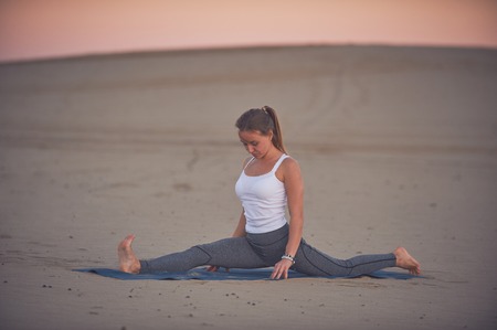 Beautiful young woman practices yoga asana Hanumasana - monkey king pose in the desert at sunset.の写真素材