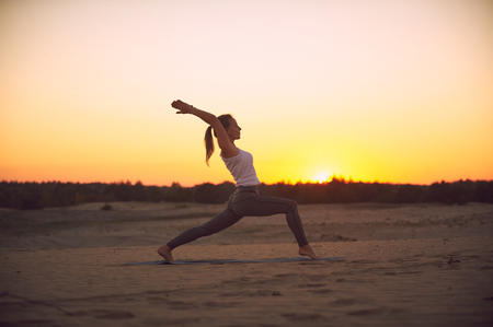 Beautiful young woman practices yoga asana Virabhadrasana 1 - warrior pose 1 in the desert at sunset.の写真素材