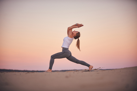 Beautiful young woman practices yoga asana Virabhadrasana 1 - warrior pose 1 in the desert at sunset.の写真素材