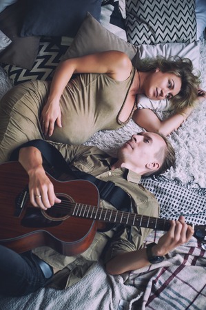 man playing a guitar and his pregnant wife are lying on a bed in a loft roomの写真素材