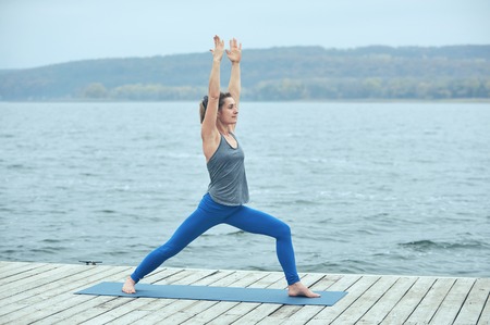 Beautiful young woman practices yoga asana Virabhadrasana 1 - warrior pose 1 on the wooden deck near the lakeの写真素材