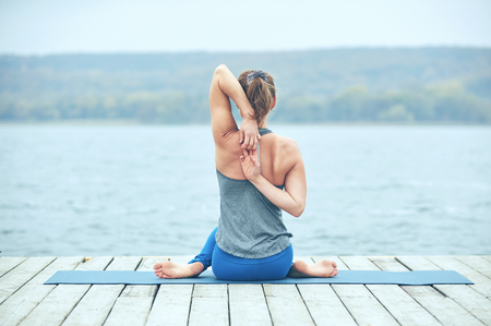 Beautiful young woman practices yoga asana Gomukhasana - Cow face pose on the wooden deck near the lakeの写真素材