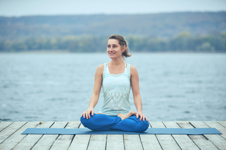 Beautiful young woman practices yoga asana Padmasana - Lotus pose on the wooden deck near the lakeの写真素材