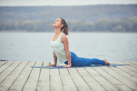 Beautiful young woman practices yoga asana upward facing dog on the wooden deck near the lake