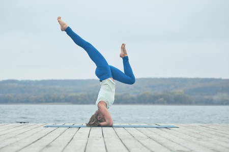 Beautiful young woman practices yoga asana Shirshasana - Headstand pose on the wooden deck near the lakeの写真素材