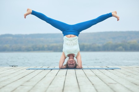 Beautiful young woman practices yoga asana Shirshasana - Headstand pose on the wooden deck near the lakeの写真素材