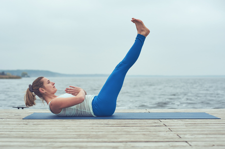 Beautiful young woman practices yoga asana Paripurna Navasana on the wooden deck near the lakeの写真素材
