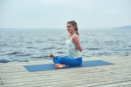 Beautiful young woman practices yoga asana Gomukhasana - Cow face pose on the wooden deck near the lakeの写真素材