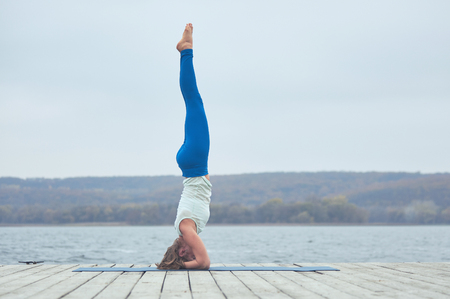 Beautiful young woman practices yoga asana Shirshasana - Headstand pose on the wooden deck near the lakeの写真素材