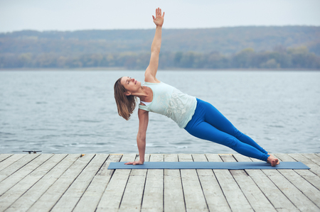 Beautiful young woman practices yoga asana Vasishthasana - side plank pose on the wooden deck near the lakeの写真素材