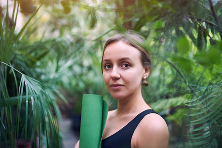 Portrait of cute young woman holding a rolled yoga mat in jungle. Sunny dayの写真素材