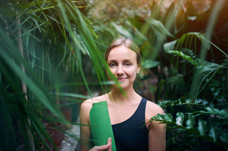 Portrait of cute young woman holding a rolled yoga mat in jungle. Sunny dayの写真素材