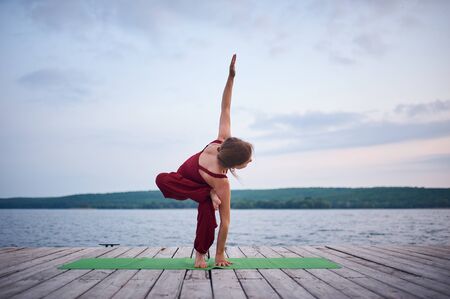 Beautiful young woman practices yoga on the wooden deck near the lakeの写真素材