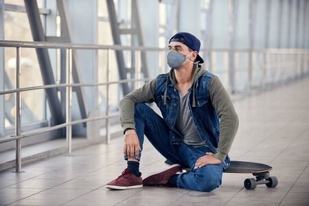 Handsome young caucasian man in hoody sitting on a skateboard on the street with a medical face mask on. Coronavirus concept photo.の写真素材