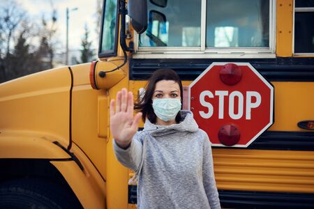 A woman in a protective mask stands on the background of a school bus. Raises a hand and stops the...