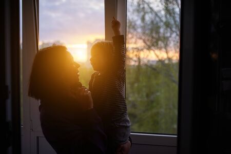 Mother with baby looks out the window at sunset in isolation at home for virus outbreak. Stay home concept.の写真素材