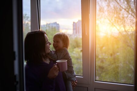 Mother with baby looks out the window at sunset in isolation at home for virus outbreak. Stay home...