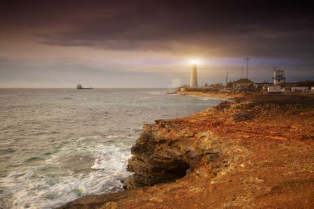 Lighthouse on a rock coast shining at sunset. large ship is seen in the sea.の写真素材