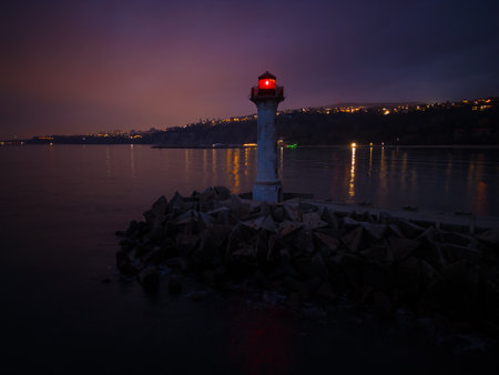 lighthouse on the seashore with red light at night against the background of the lights of the ships