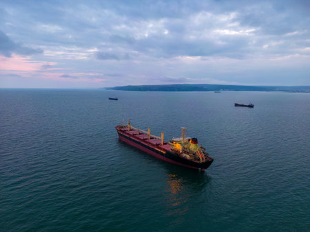aerial view of a bulk carrier at evening showcases a stunning display of lights reflecting off the water as the massive vessel cuts through the waves. The contrast between the darkness of the ocean and the brightly lit ship creates a captivating and atmospheric scene.の写真素材