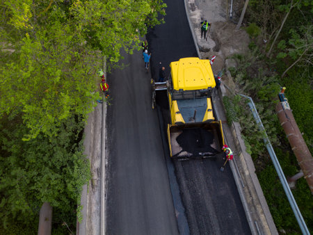 Aerial view of the construction of a new road as asphalt pavers, rollers, machinery, and workers...