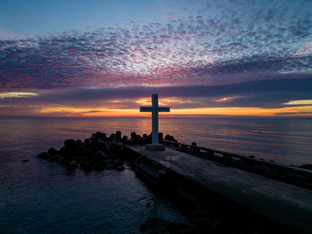 A flight around a Christian Holy cross early in the morning at sunrise. The large cross stands on the edge of a breakwater on the sea coastの写真素材
