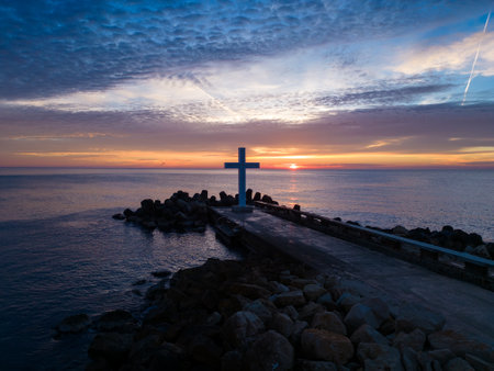 A flight around a Christian Holy cross early in the morning at sunrise. The large cross stands on the edge of a breakwater on the sea coastの写真素材