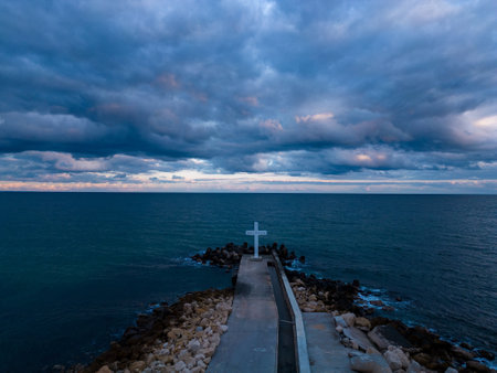 A large Christian cross stands at the edge of a pier against a dramatic sky and sea, seen from aboveの写真素材