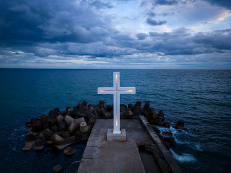 A large Christian cross stands at the edge of a pier against a dramatic sky and sea, seen from aboveの写真素材
