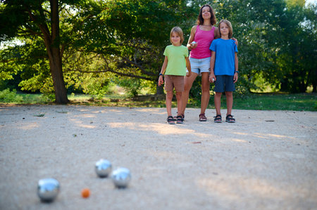 A family plays petanque in a sunny park. The mother watches as her two sons throw metal balls towards the target, enjoying a casual outdoor activity togetherの写真素材