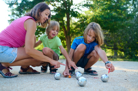A mother and her two sons measure the distance between petanque balls and the target ball. A fun moment of focus and teamwork during an outdoor family game in a sunny parkの写真素材