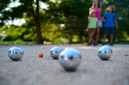 A family plays petanque in a sunny park. The mother watches as her two sons throw metal balls towards the target, enjoying a casual outdoor activity togetherの写真素材