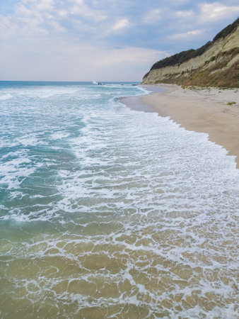A tranquil coastal scene with turquoise waves gently washing onto a sandy beach, bordered by rugged cliffs. Perfect for nature, travel, or seascape photography themesの写真素材