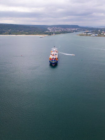A cargo container ship navigates the harbor under a cloudy sky while a small boat moves nearby towards the shoreline at middayの写真素材