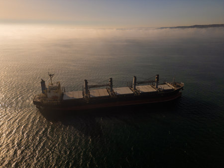 Aerial drone footage of a cargo ship sailing on misty waters with sunlight reflections, creating a calm and mysterious maritime scene, perfect for industrial and nautical themesの写真素材