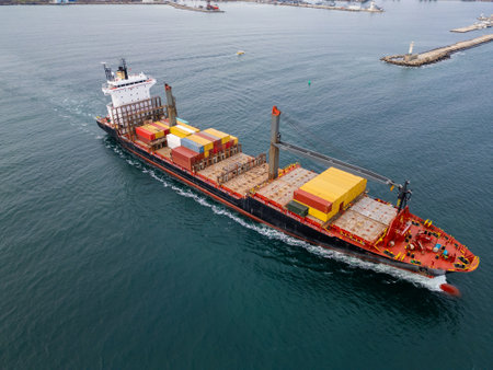 Aerial drone view of a red cargo ship loaded with containers, sailing through the Black Sea near the port of Varna, Bulgaria. Industrial scenery and cranes create a dynamic backdrop for maritime tradeの写真素材